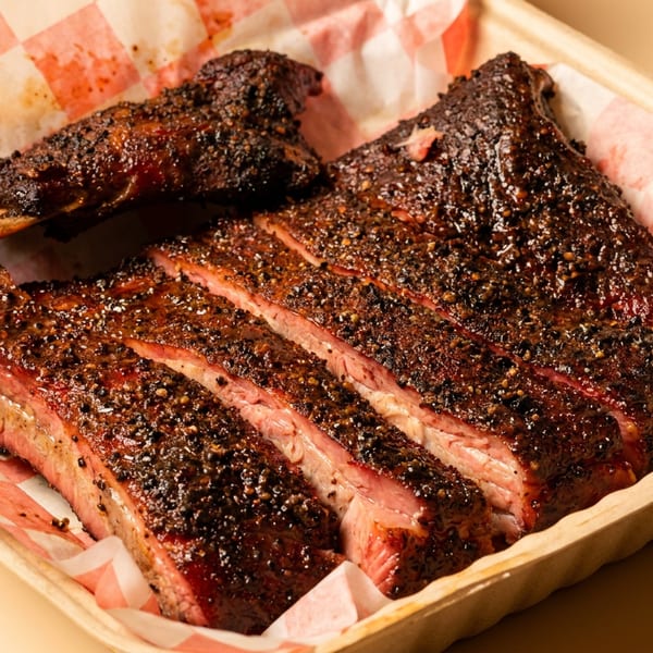 Sliced, dark-crusted smoked ribs in a disposable container with red and white checkered paper.