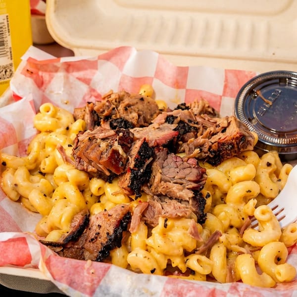 Macaroni and cheese topped with pulled barbecue meat, served in a disposable container with sauce and a fork.